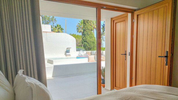 A bedroom with a bed, a wooden door, and a view of an outdoor area with a pool and plants. - Two Bedroom Villa (Porches)