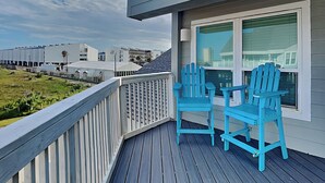 A balcony with two blue Adirondack chairs, a view of a pier, and a building with a white facade in the background. - Fantasea Cove by Silver Sands Vacation Rentals (Port Aransas)