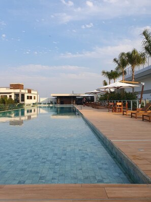 Una piscina con suelo de baldosas, rodeada de sillas de descanso y sombrillas, y edificios al fondo.