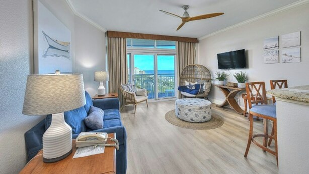 A spacious living room with a blue sofa, a wooden coffee table, a round ottoman, a ceiling fan, and a view of the ocean through large windows. - Horizon PH2 -2BR/2BA -Myrtle Beach -I/O Pools (Myrtle Beach)