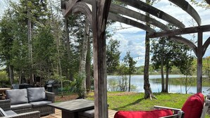 Una terraza exterior con mobiliario de mimbre y una pérgola de madera con vistas a un lago.