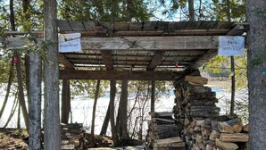 Una estructura de madera rústica con letreros que indican 'Bois extérieur' y 'Bois intérieur' para áreas exteriores e interiores, respectivamente, rodeada de árboles y una masa de agua.