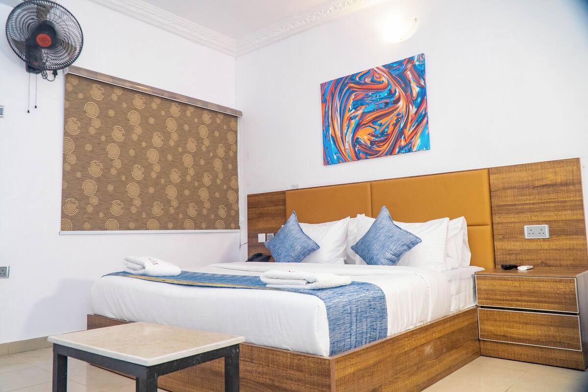 A hotel room with a large bed, a bedside table, a wall-mounted fan, and a colorful abstract painting.