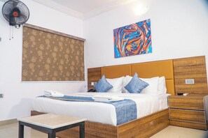 Habitación de hotel con una cama grande, una mesita de noche, un ventilador de pared y un cuadro abstracto de colores.