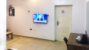 A hotel room with a television displaying a news channel, a white door, a small table with a coffee maker, and a chair. - Ringruby Hotel, United Estate (Lekki)