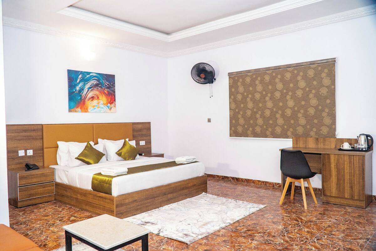 A hotel room with a bed, bedside table, desk, chair, and artwork on the wall.
