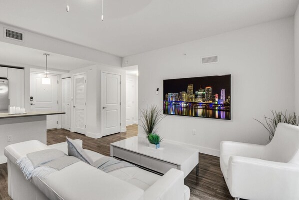 A modern living room with a white sofa, a coffee table, and a cityscape painting on the wall. - Comfort & Convenience: 2BR Unit with Free Parking! (Miami)