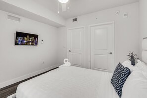 A bedroom with a large bed, two white doors, a wall-mounted TV displaying a cityscape, and a small plant on the nightstand. - Comfort & Convenience: 2BR Unit with Free Parking! (Miami)