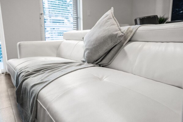 A white leather sofa with a grey throw blanket and pillow in a modern living room with a window and blinds.