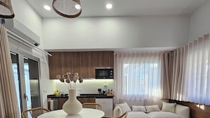 A modern living room with a dining area, a kitchen in the background, and a large window with curtains.
