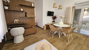 A modern living room with a wooden wall unit, a white sofa, a dining table with wicker chairs, and a flat-screen TV mounted on the wall.