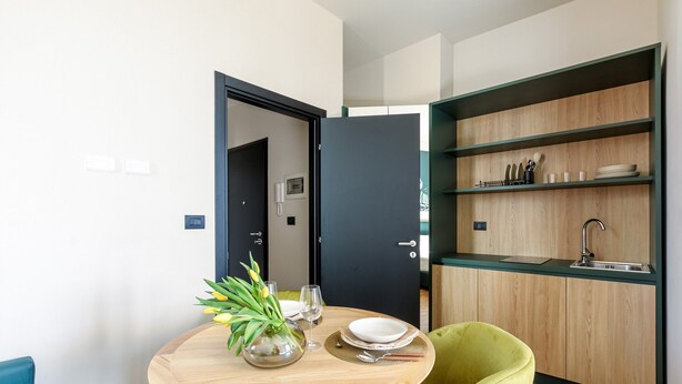 A compact dining area with a round table set for two, a vase with yellow flowers, and a green chair. A dark door leads to a hallway with a phone and a light switch. - Cozy and Central Studio in Bovisa, Milan (Milan)