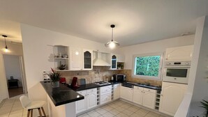 A modern kitchen with white cabinets, a black countertop, and a gas stove. There are two white bar stools and a window with a view of greenery. - Apartment in Carantec Near Sandy Beaches (Carantec)