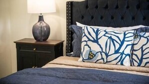 A bedroom with a dark upholstered headboard, a bedside table with a lamp, and a bed with a blue blanket and patterned pillows. - The Ace, 1BR King Duplex by Velvet Ditch Villas (Oxford)