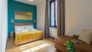 Une chambre d’hôtel moderne comprenant un lit, un canapé, une fenêtre avec des rideaux, une œuvre d’art murale et un vase avec des fleurs.