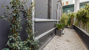 Un étroit sentier pavé de briques, bordé d’un immeuble gris foncé aux fenêtres grillagées et d’une petite plante en pot.