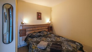 A bedroom with a wooden headboard, a bed with patterned bedding, a nightstand with a lamp, and a wall-mounted mirror. - T2 cosy - Vue Golf - Proche Plage (Pornic)