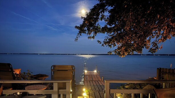 Una terraza de madera con vista a una masa de agua tranquila al anochecer, un muelle que se adentra en el agua y mobiliario exterior.