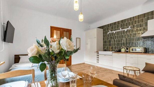 Une cuisine moderne avec des meubles blancs et un pare-feu vert, un coin repas avec une table en bois dressée pour deux personnes, et un salon confortable avec un canapé marron et un vase de fleurs.