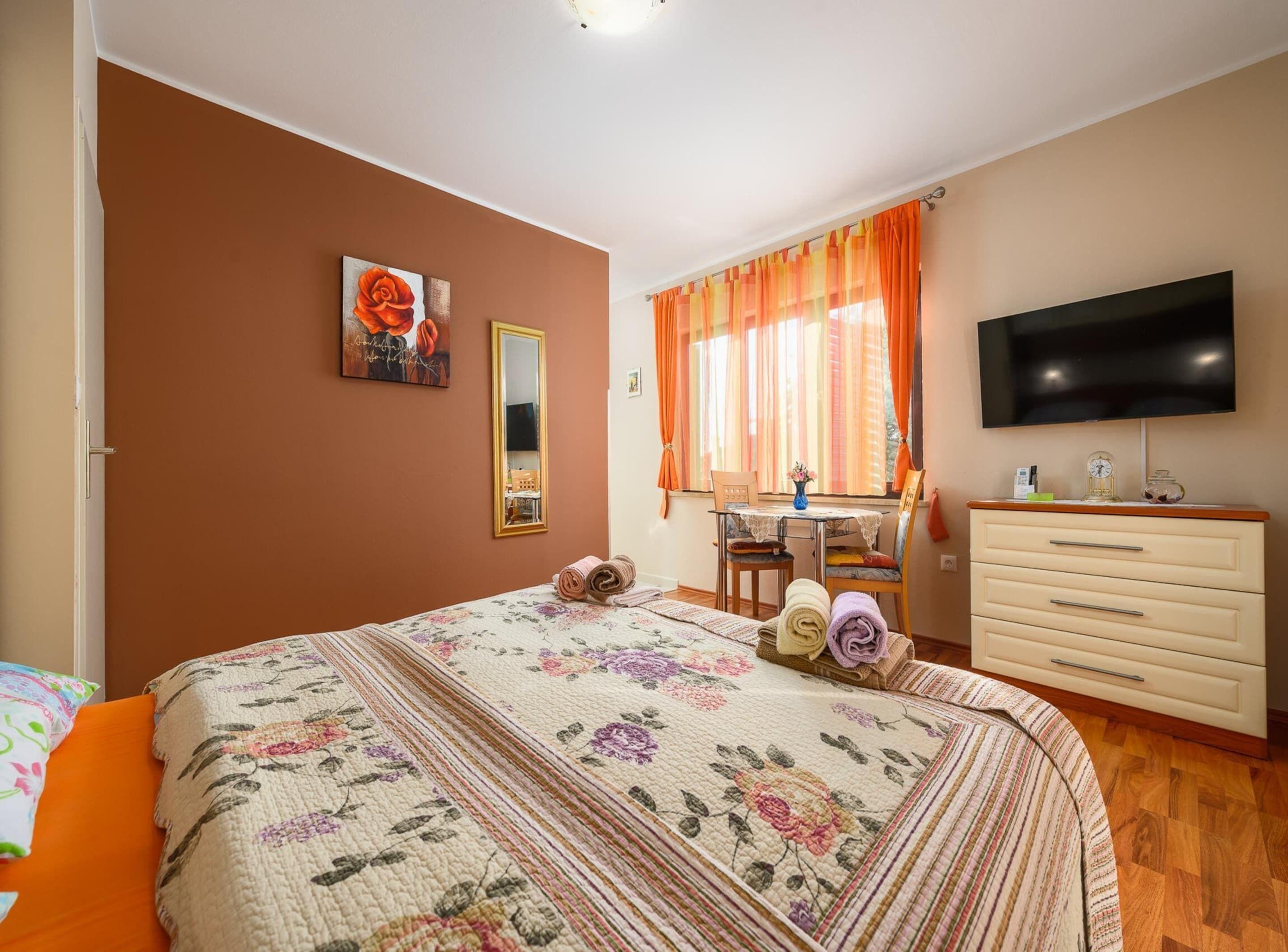 A bedroom with a bed, a desk, a dresser, a TV, and a window with curtains.