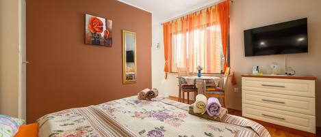 A bedroom with a bed, a desk, a dresser, a TV, and a window with curtains.