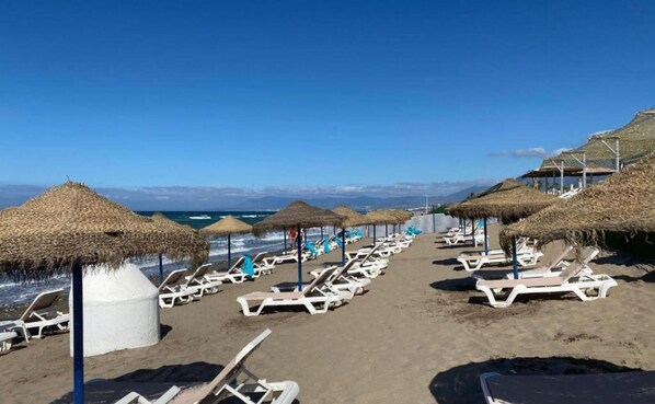 Una playa con filas de tumbonas y sombrillas de paja, cielo despejado y vistas al océano y montañas distantes.
