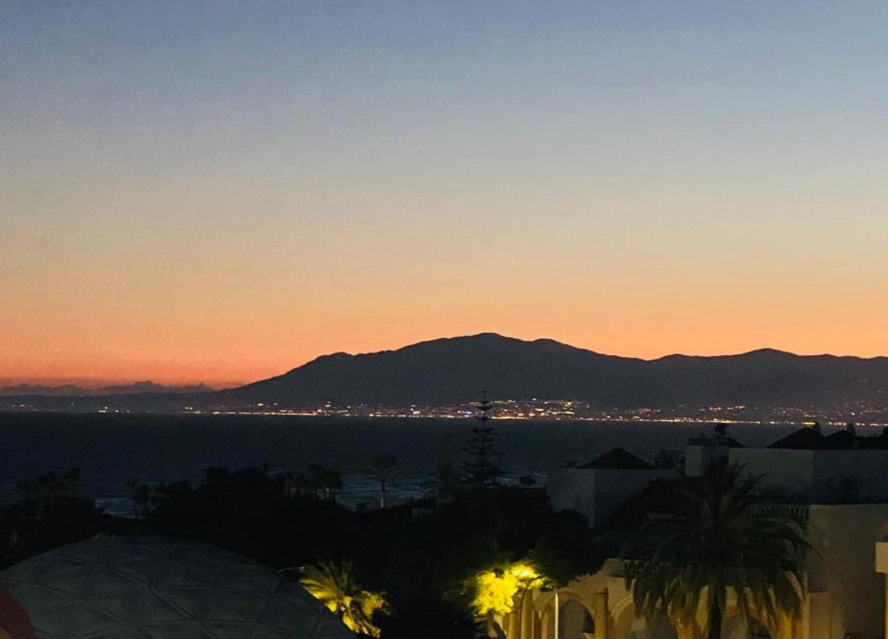 A coastal cityscape at dusk with mountains in the background and a view of the sea.
