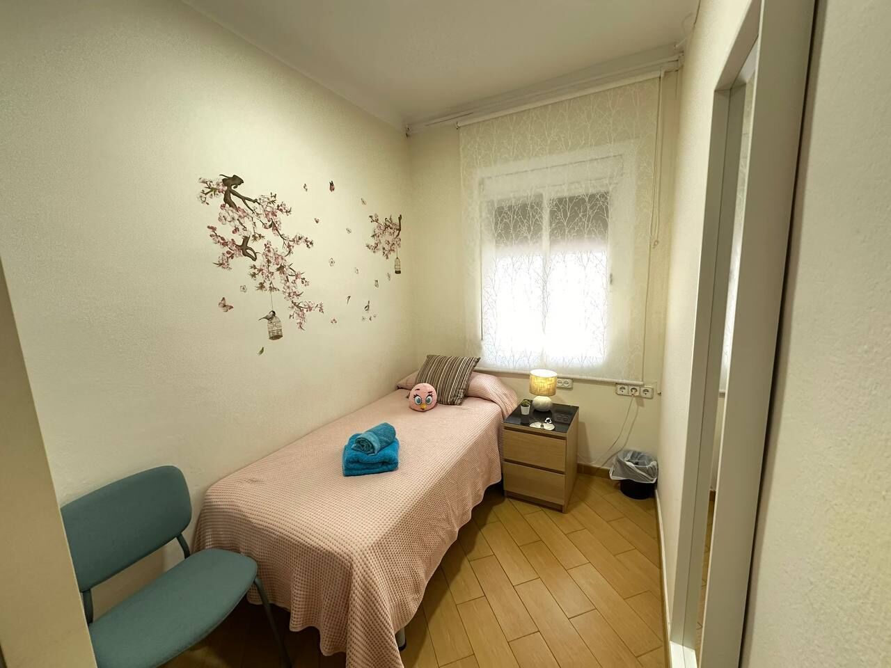 Un dormitorio pequeño y ordenado con una cama individual, una mesita de noche, una silla y una ventana con cortinas de gasa.
