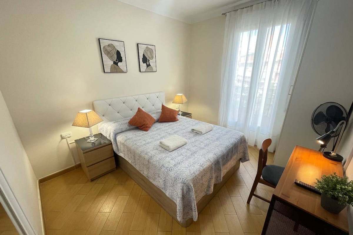 Une chambre à coucher comprenant un lit, deux tables de chevet, un bureau avec une chaise, un ventilateur et une fenêtre avec des rideaux.