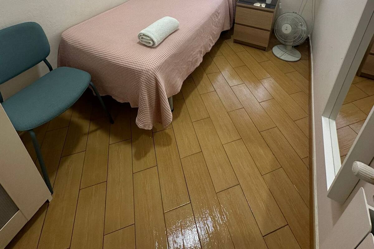 Une petite chambre avec un lit simple, une chaise, une table de chevet avec une lampe et une fenêtre donnant sur des arbres sans feuilles.