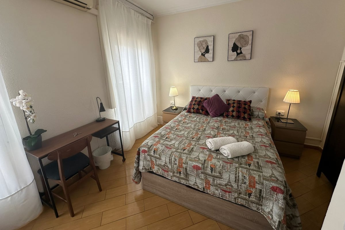 Une chambre d’hôtel bien rangée, avec un lit, deux lampes de chevet, un bureau avec une chaise, une petite table et une plante en pot.