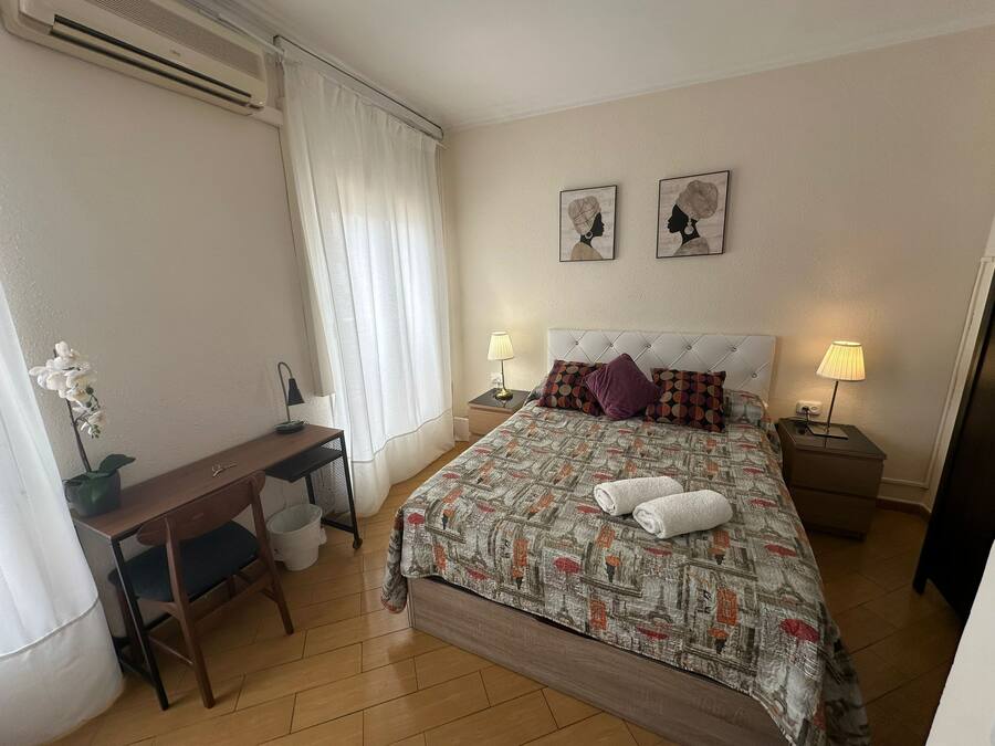 Habitación de hotel ordenada con cama, dos lámparas de noche, un escritorio con silla, una mesita y una planta en maceta.