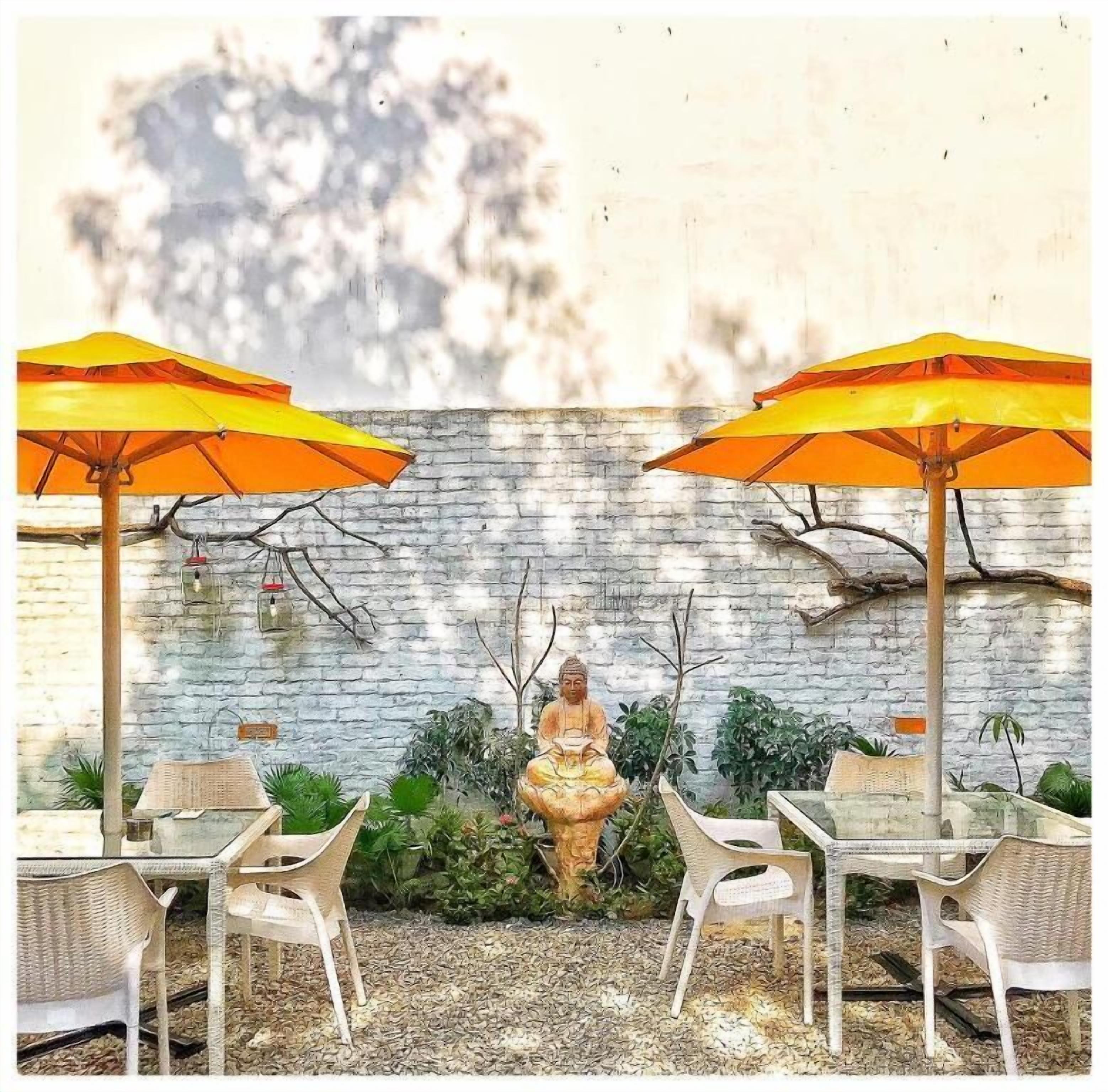 A patio with two wicker chairs and two orange umbrellas, a Buddha statue, and a stone wall with climbing plants.