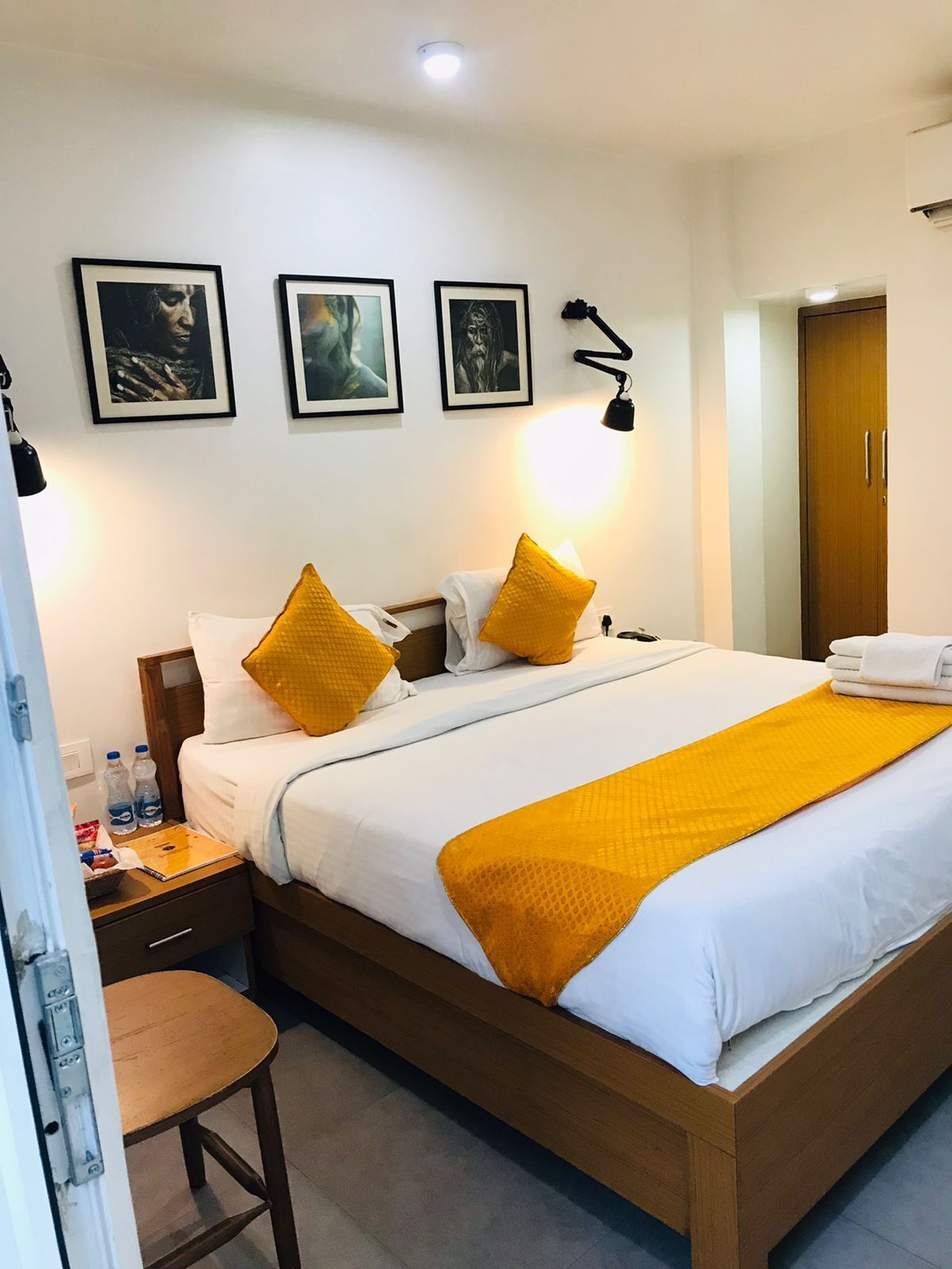 A hotel room with a wooden bed, white bedding, and two yellow pillows. There are framed pictures on the wall, a nightstand with a lamp, and a wooden chair.