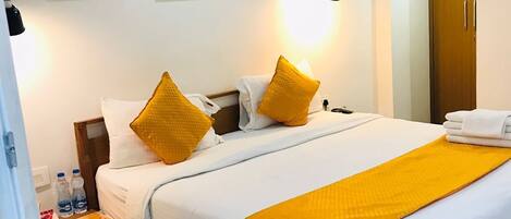 A hotel room with a wooden bed, white bedding, and two yellow pillows. There are framed pictures on the wall, a nightstand with a lamp, and a wooden chair.