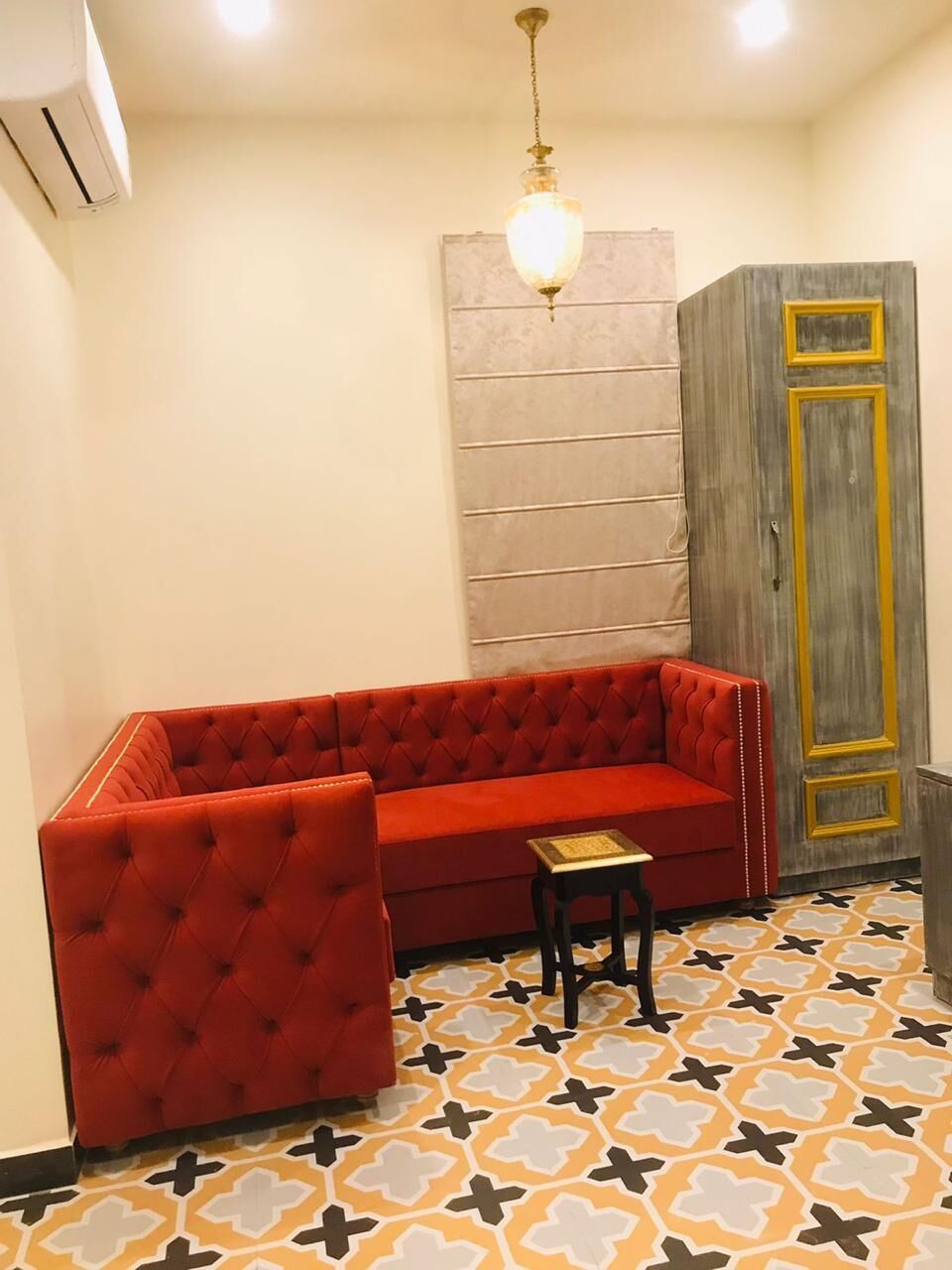 Un sofá rojo, un taburete pequeño, un suelo estampado, un aire acondicionado instalado en la pared y un armario de madera con detalles dorados.