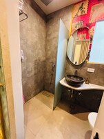 Un bagno con pavimento piastrellato color beige, un lavandino bianco con una ampia vasca, uno specchio e una doccia con porta.