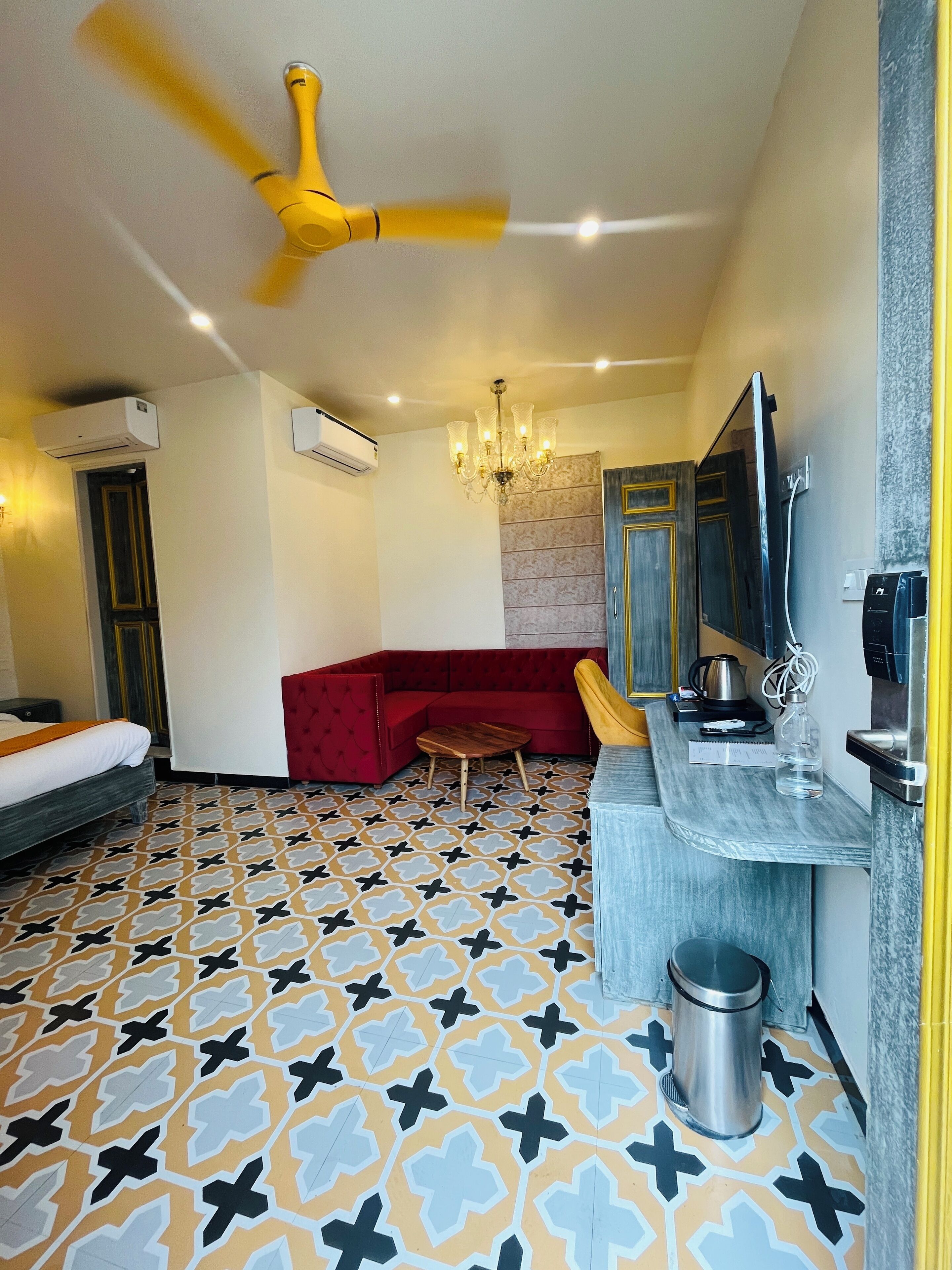 A modern hotel room with a patterned floor, a red sofa, a small wooden table, a ceiling fan, an air conditioning unit, a chandelier, a mirror, a sink, and a trash bin.