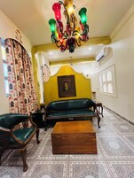 A room with a green sofa, two wooden chairs, a wooden coffee table, a patterned curtain, a chandelier, and a wall-mounted air conditioner.
