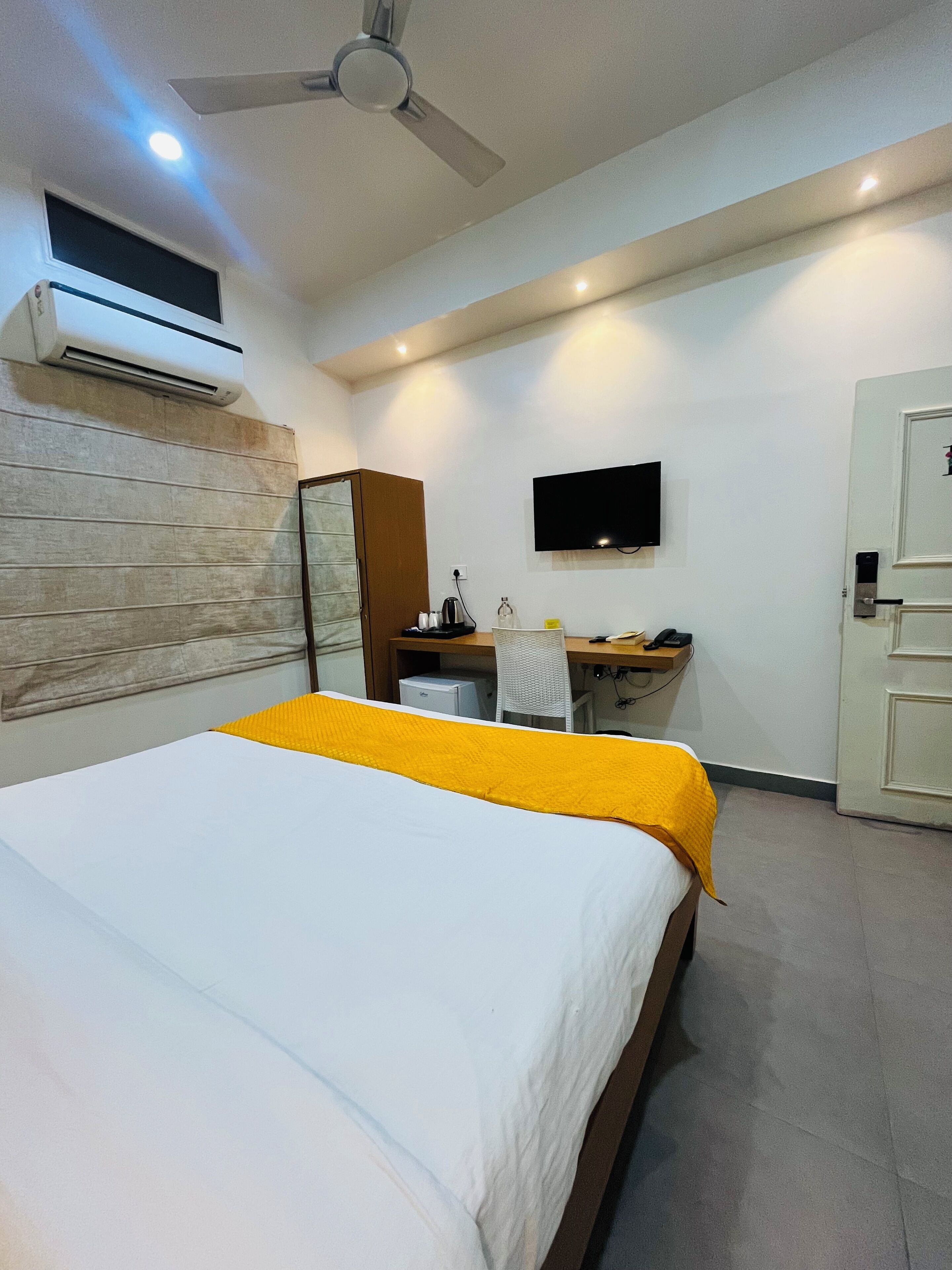 A hotel room with a bed, a desk, a chair, a TV, and an air conditioning unit.