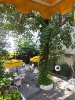 An outdoor seating area with white chairs and yellow umbrellas, surrounded by greenery and a tree.