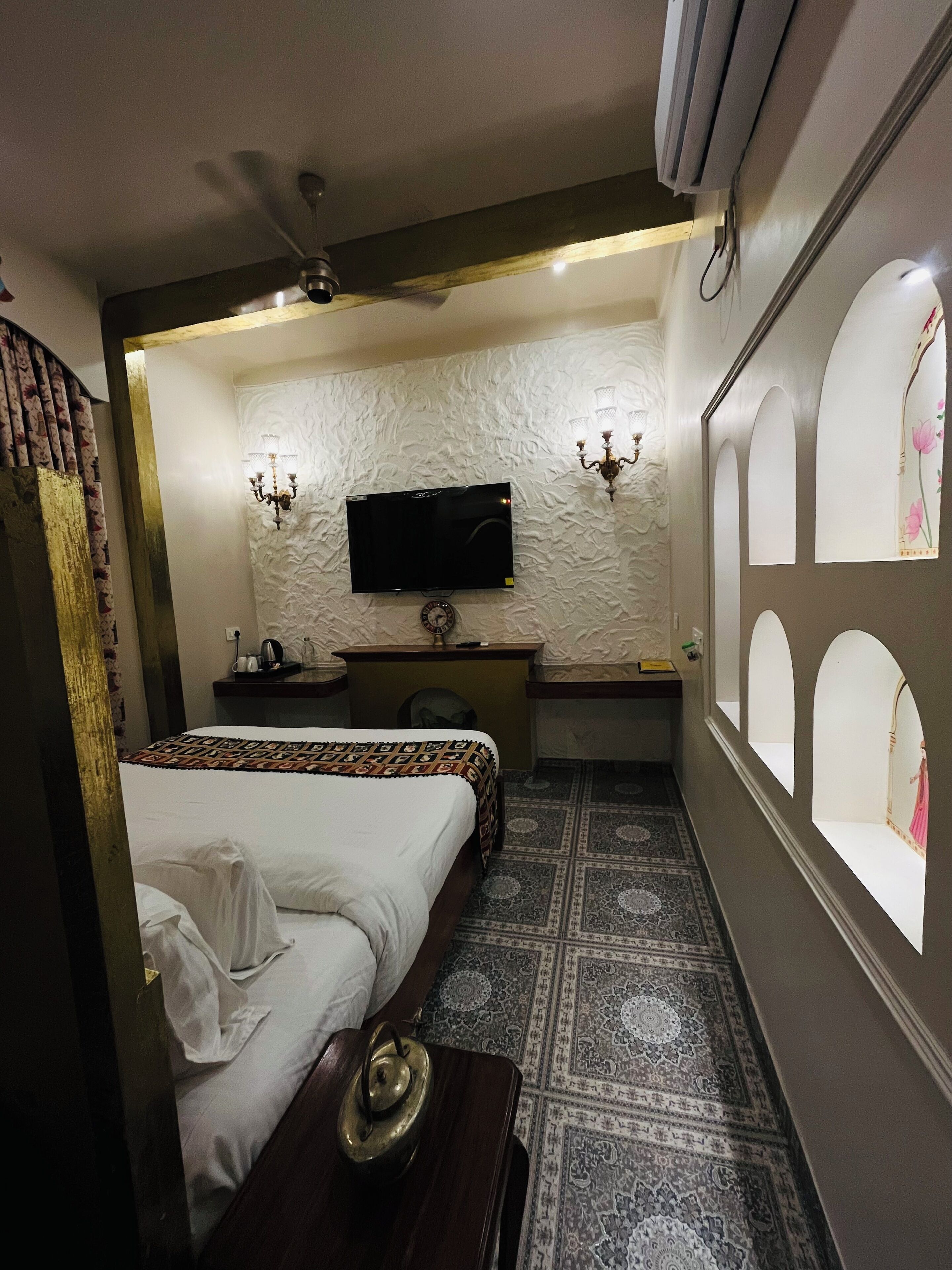 A hotel room with a bed, a desk, a television, a clock, and a patterned rug.