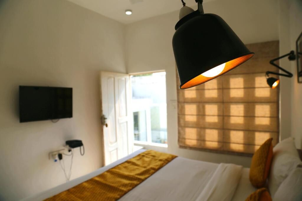Habitación de hotel con cama, televisión montada en la pared, ventana con persianas y lámpara colgante.