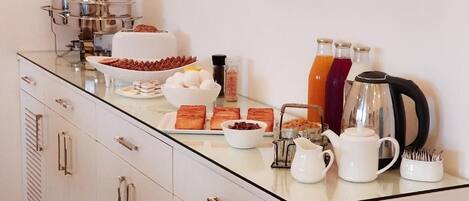 Ein Frühstücksbuffet mit verschiedenen Speisen, einer Kaffeemaschine und einem Wasserkocher auf einer Glastheke.