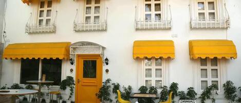 A building with multiple windows, some with yellow awnings, and several outdoor tables and chairs.