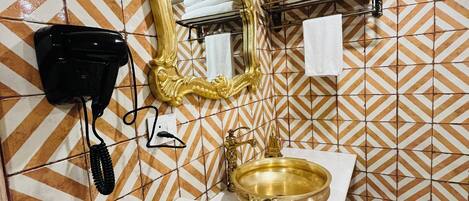 A bathroom with a patterned tile wall, a gold-framed mirror, a sink with a gold basin, a black hairdryer, and a towel rack.