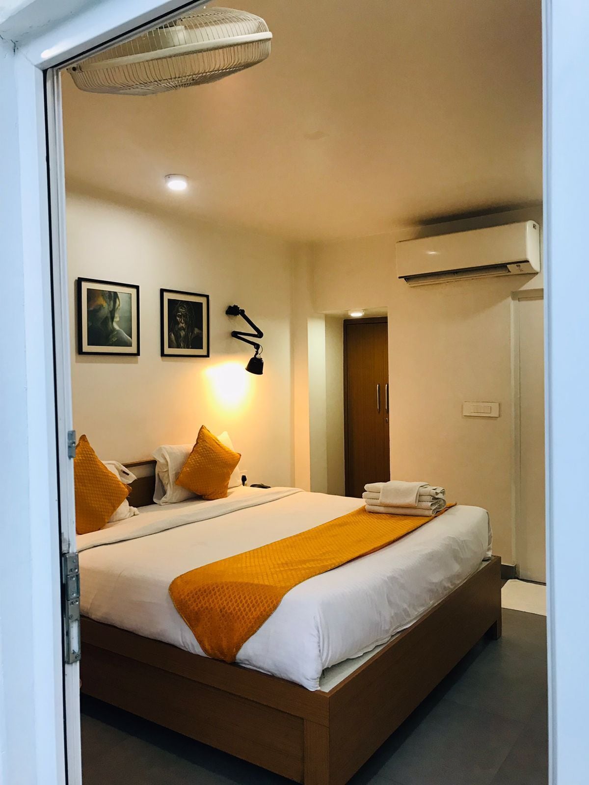 A hotel room with a large bed, a bedside table, a lamp, two framed pictures on the wall, an air conditioning unit, and a door.