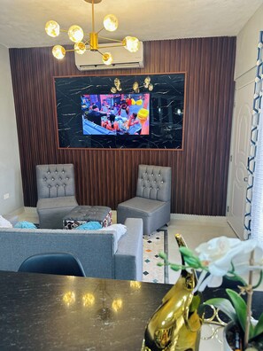 A living room with a flat-screen TV mounted on a wall, two grey upholstered chairs, a small coffee table, and a decorative plant.