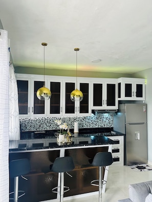 A kitchen with white cabinets, a black countertop, two hanging pendant lights, a refrigerator, and a bar area with stools.