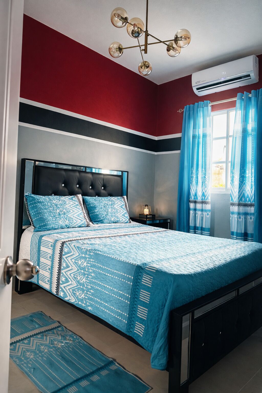 A bedroom with a bed, a nightstand, a lamp, a window with curtains, and an air conditioning unit.